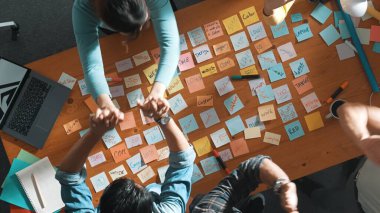 Top view of business team giving high five to celebrate project while standing at table with sticky notes and laptop. Aerial view of designer clapping hands to congratulate successful idea. Symposium.