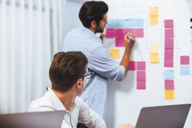 Çeşitli ofis çalışanlarından oluşan bir grup, toplantı odasındaki beyaz tahtaya yapışkan bir notla stratejik iş pazarlama planlaması üzerine beyin fırtınası yapıyor. İş yerinde verimli bir takım çalışması. Tedbirli