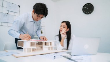 Profesyonel Asyalı erkek mimar ev modelinin uzunluğunu ölçmek için cetvel kullanırken, beyaz tenli genç meslektaşı ev modeliyle toplantı masasındaki verileri analiz etmek için dizüstü bilgisayar kullanıyor. Tertemiz..