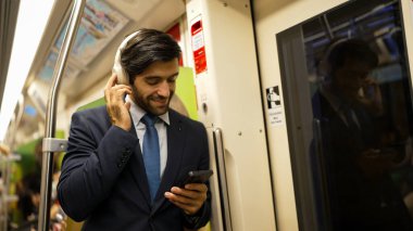 Akıllı yönetici trende dururken kulaklık takar ve müzik dinler. Profesyonel iş adamı, şarkıları dinlerken ve şarkı seçerken bölüme yaslanıyor. İş konsepti. Sevinçli..