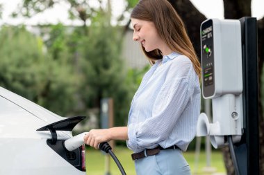 Açık yeşil şehir parkı manzaralı EV şarj istasyonundan genç kadın EV elektrikli araçları şarj ediyor. Çevre dostu şehir ulaşımı ve çevre dostu EV araba seyahati. Yükselt