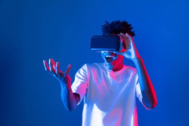 Sanal gerçekliği dijital fütüristik teknolojiye bağlayan pembe mavi neon duvarda VR 'dan bakan genç Afrikalı Amerikalı' ya şaşırdım. Entrika.