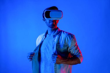 VR gözlüklü beyaz bir adam neon arka planda günlük kıyafetleri ayarlıyor. Gülümseyen genç teknolojik yenilik simülasyonu kullanarak metaevren ya da sanal dünyaya girerken ayakta duruyor. Sapma.