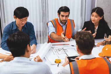 Mimari proje üzerinde birlikte çalışan çeşitli inşaat mühendisi ve müvekkil grupları, inşaat planını gözden geçirip toplantı masasında plan oluşturuyorlar. Tedbirli