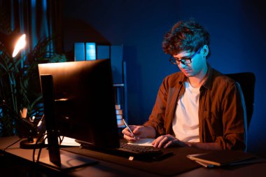 Genç işadamı, Neon Light Modern Ofis 'teki yeni proje surround çalışma masası için kağıt üzerinde tasarım düşünüyor. E-posta ile hediye göndermeden önce not defterine yeni bir iş düşünüyorum. Toplayıcı.