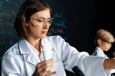 Kimya bilimini öğrenen öğretmen yardımcısı, laboratuarda deney yapıyor. Laboratuvar önlüğü ve STEM sınıfında güvenlik gözlüğü takıyor. Öğretmen masadaki test tüpünü kendine güvenerek idare ediyor. Etkinlik.