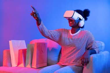 VR 'lı genç Afrikalıların alışverişinin tadını çıkarın. İnternette hologram uygulaması, güzellik ürünleri, neon ışıklandırma, oturma odasındaki koltukta alışveriş torbası bulunan canlı dijital fütüristik teknoloji. Entrika.