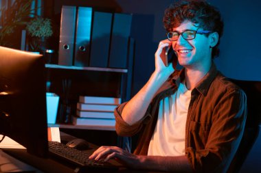 Genç iş adamı akıllı telefondan müşteriyi ya da iş arkadaşını arıyor, neon modern ofisteki yaratıcı sunum surround çalışma masasının bilgisayar ekranına bakıyor. Yeni proje kavramı. Toplayıcı.