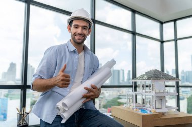 Modern ev projesini düşünen profesyonel mimar mühendisinin portresi güvenlik kaskı ve günlük kıyafetler giyerken. Akıllı iç mimar ev modelinin yanında proje planları tutuyor. İzleme.