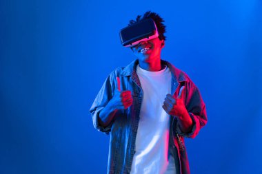 VR kullanan genç Afro-Amerikalı, boş uzay ürünlerini işaret ederek metaevren dünyasındaki mavi, neon ışıklı duvar fütüristik teknolojisi hakkında detaylı bilgi veriyor. Entrika.