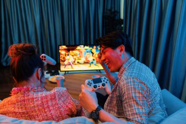 joystickli bir çift, TV ekranında dövüş oyunu oynuyor, erkek, kız arkadaşıyla yarışma seviyesini geri kazanıyor, modern evdeki neon ışıklı oturma odasında kanepeye oturuyor. İnfobahn.