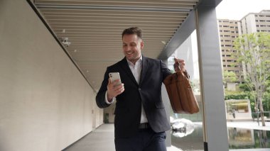 Happy proje yöneticisi koridorda yürürken cep telefonunu kontrol ediyor. Profesyonel iş adamı başarılı projesini kutlarken ya da satışları artırırken takım elbise giyer ve bavulu toplantı için tutar. Urbane.