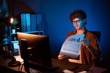 Genç işadamı, Neon Light Modern Ofis 'teki yeni proje surround çalışma masası için kağıt üzerinde tasarım düşünüyor. E-posta ile hediye göndermeden önce not defterine yeni bir iş düşünüyorum. Toplayıcı.