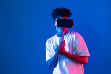 VR kullanan genç Afrikalı Amerikalı oyuncu sanal gerçeklik dünyasını dijital fütüristik teknolojiye bağlayan mavi neon ışıklandırma duvarında 3 boyutlu sanal silah avcısını kullanıyor. Entrika.