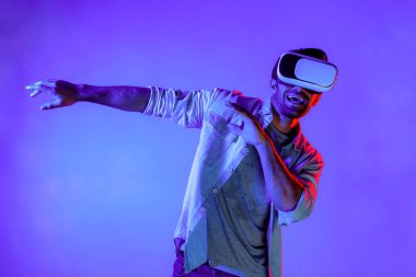 Yakışıklı, dans eden ve müziğe hareket eden bir adam. VR gözlüğü ve neon ışıklı arka plan kullanarak. Beyaz erkek oyuncu, teknolojik yenilikler giyerek pop müzik dinlerken ayakta duruyor. Sapma.