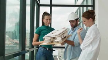 Proje yöneticisi ev modelini elinde tutarken, mimar mühendis takımı da ev tasarımı veya bina inşaatı hakkında fikir alışverişinde bulunuyor. Şehrin penceresinin yanında iç mimar duruyor. İzleme
