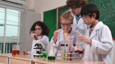 Beyaz çocuk kimyasal çözeltiyi örneğin çeşitli öğrenciler deney yaparken bırakıyor. Amerikalı bir kız, öğretmeniyle birlikte tahtada çocuklara bakarken renkli sıvıyı inceliyor. Pedagoji.