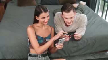 Genç çift, kredi kartıyla ödemek için internet üzerinden ödeme uygulaması ve dijital cüzdan kullanarak yatak odasında oturuyor. Mobil internet üzerinden alışveriş ve modern alımlar. Düzenle