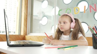 Tatlı akıllı kız sınıf çalışması yapıyor ya da öğretmenini dinlerken internetten öğreniyor. Mutlu öğrenci yazıları, çizimleri, bilgisayar ve renkli kalemlerle kulaklık takarak kağıt üzerinde çalışması. Etkinlik.
