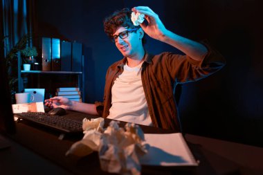 Aşırı çalışan projeyle uğraşan stresli yaratıcı yönetici buruşmuş kağıtların etrafını sararken gece vakti modern ofisteki gündelik gömlekle dizüstü bilgisayar ekranına e-posta göndermeyi bekliyor. Toplayıcı.