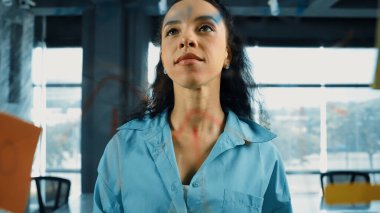 Pazarlama fikrini düşünen yetenekli, genç ve güzel İspanyol proje yöneticisinin portresi. Ofisteki cam tahtada yapışkan notlar kullanarak iş planı yapan profesyonel bir iş kadını. Yönlendirici.