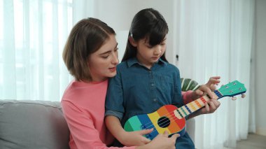 Kafkasyalı bir anne evde akustik müzik öğretirken ukulele çalan mutlu kız. Sevimli çocuk enstrüman öğreniyor. Kafkasyalı mutlu anne ve kız birlikte vakit geçiriyorlar. Pedagoji.