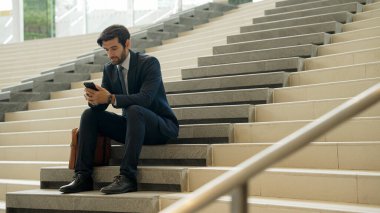 Akıllı proje yöneticisi merdivenlerde otururken cep telefonuna bakıyor. Çekici Kafkasyalı iş adamı telefon kullanarak çalışıyor. Profesyonel yatırımcı iş stratejisi planlamak için telefonu kullanıyor. Sevinçli..