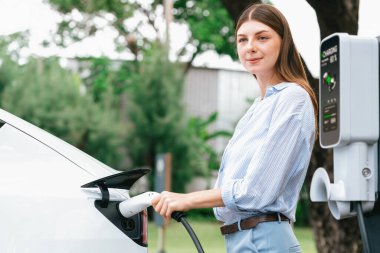 Açık yeşil şehir parkı manzaralı EV şarj istasyonundan genç kadın EV elektrikli araçları şarj ediyor. Çevre dostu şehir ulaşımı ve çevre dostu EV araba seyahati. Yükselt