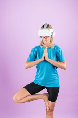 Pembe arka planda gömlek giyerken yoga yapan beyaz bir kız. Mutlu akıllı kadın pembe stüdyoda dikilirken yoga yapıyor ve sanal gerçeklik gözlüğü ya da görsel gerçeklik kulaklığı kullanıyor. Kısıtlanma.