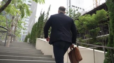 Yeşil şehirde bavulunu tutarken resmi takım elbiseli beyaz bir müdürün merdivenlerden çıkışının arka görüntüsü. Profesyonel bir iş adamı iş yerine giderken aynı zamanda Eko Şehri 'nde merdivenleri çıkıyor. Urbane..