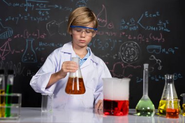 Laboratuvardaki sarışın çocuk laboratuvar önlüğü giyer ve STEM sınıfında kimya bilimi öğrenir. Öğrenci portakal suyu ile deney kabını tutuyor. Masaya bir sürü şişe koy. Etkinlik.