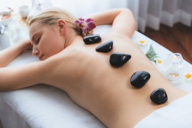 Lüks tatil köyündeki kaplıcalarda sıcak taş masajı. Gün ışığı sükunet ortamında, mutlu kadın müşteri bazalt taş masajından zevk alıyor. Rahatlatıcı bir sıcaklıkla vücudun üzerinde süzülüyor. Sessiz.