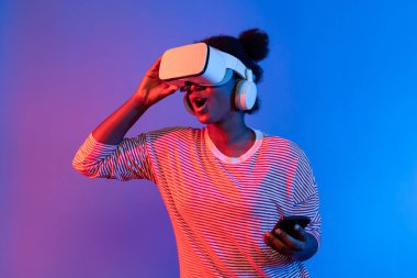 VR kulaklıklı ve akıllı telefonlu genç Afrikalı kadın canlı neon ışıklandırma arka planı sanal gerçekliği ve mobil bağlantı öğrenme teknolojisiyle metaevrene doğru yol alıyor. Entrika.