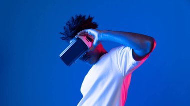 Sanal gerçekliği dijital fütüristik teknolojiye bağlayan pembe mavi neon duvarda VR 'dan bakan genç Afrikalı Amerikalı' ya şaşırdım. Entrika.