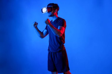VR gözlüklü mutlu adam neon ışıklı arka planda egzersiz yapıyor ve kolunu esnetiyor. Günlük kıyafetleri olan beyaz bir insan sanal gerçeklik simülatörü teknolojisi kullanarak rahatlıyor ve egzersiz yapıyor. Sapma.