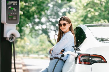 Yolculuk sırasında doğal orman ya da milli parktaki elektrikli araba için şarj olan genç bir kadın. Tatil ve tatil boyunca çevre dostu seyahat. Yükselt