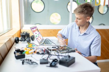 Mavi tişörtlü sarışın okul çocuğu dizüstü bilgisayarda kod izliyor ve STEM sınıfında tornavida kullanıyor. Dizüstü bilgisayar, elektrik kablosu, şarj aleti ve dizüstü bilgisayar. Öğretim.