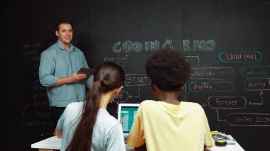 Kafkasyalı öğretmen elinde tablet tutarken öğrenci sorusuna cevap veriyor. Beyaz kız, BTMM teknoloji sınıfında dizüstü bilgisayar ekranı görüntülerken kod ile tahtaya işaret ediyor. Öğretim.
