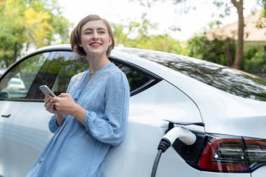 Yeşil kırsal kesimde tatilde güzel bir genç kadın elektrikli araçla şarj olurken akü durumunu kontrol ediyor. Çevre dostu seyahat zekası. Kalıcı.