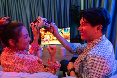 joystick 'in çaprazına sahip bir çift oyuncu arka tarafta televizyon ekranında kazananla kavga oyunu oynuyor modern rahat evinde neon ışıklı oturma odasında kanepesinde oturuyor. İnfobahn.