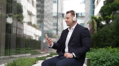 Kafkasyalı iş adamı cep telefonunu tutarken rahatlatıcı müzik dinliyor. Yetenekli mutlu yönetici müzik açar ve yeşil şehir veya parkta otururken neşeli bir şekilde şarkıya geçer. Urbane.