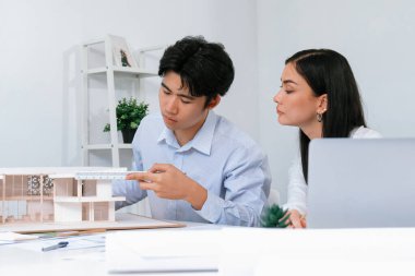 Profesyonel Asyalı erkek mimar ev modelinin uzunluğunu ölçmek için cetvel kullanırken, beyaz tenli genç meslektaşı ev modeliyle toplantı masasındaki verileri analiz etmek için dizüstü bilgisayar kullanıyor. Tertemiz..