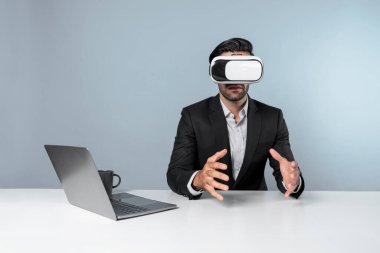 Profesyonel proje yöneticisi dizüstü bilgisayar başında otururken VR gözlüğü kullanıyor. Görsel gerçeklik kulaklığı takan yetenekli bir iş adamı metaevren ya da görsel dünyayı birbirine bağlıyor. Yenilik. Sapma.