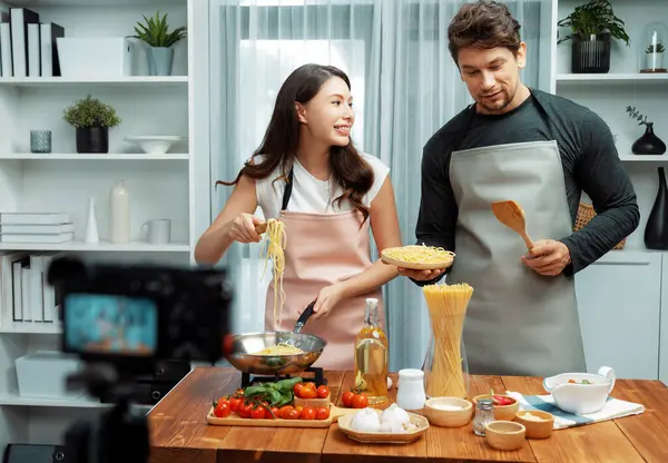 Stock image Couple chef influencers cooking special homemade of spaghetti with tongs taking to frying pan, putting seasoning and tasty sauce to make good flavor, recording on camera with live chanel. Postulate.