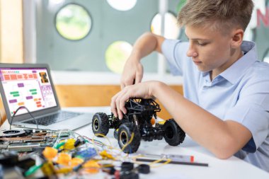 Sarı saçlı okul çocuğu, BTMM sınıfında robotik araç tamir eden bir tornavida kullanıyor. Masaya dizüstü bilgisayar, kumanda, elektrik kablosu, şarj aleti ve robot araç koyun. Öğretim.
