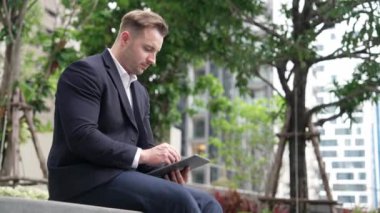 Tablet kalemle yazarken parkta oturan işadamının yan görüntüsü. Akıllı proje yöneticisinin finansal planı düşünürken veri analizine baktığı düşük açılı kamera. Urbane