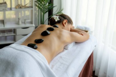 Lüks tatil köyündeki kaplıcalarda sıcak taş masajı. Gün ışığı sükunet ortamında, mutlu kadın müşteri bazalt taş masajından zevk alıyor. Rahatlatıcı bir sıcaklıkla vücudun üzerinde süzülüyor. Sessiz.