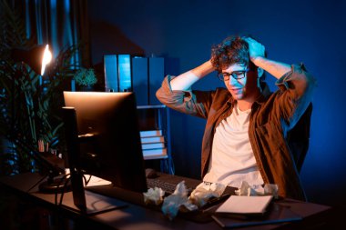 Aşırı çalışan projeyle uğraşan stresli yaratıcı yönetici buruşmuş kağıtların etrafını sararken gece vakti modern ofisteki gündelik gömlekle dizüstü bilgisayar ekranına e-posta göndermeyi bekliyor. Toplayıcı.