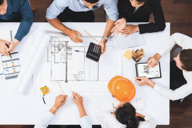 Mimari proje üzerinde birlikte çalışan, inşaat planını gözden geçiren ve toplantı masasında plan oluşturan çeşitli inşaat mühendisi ve müşteri gruplarından oluşan üst düzey bir pankart. Tedbirli