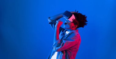 VR gözlükleri kullanarak genç Afrikalı Amerikalı şarkıcı sanal gerçeklik fantezi teknolojisinde mavi ışıklı arka planda karaoke meta uygulama ile şarkı söylemekten zevk alır. Entrika.